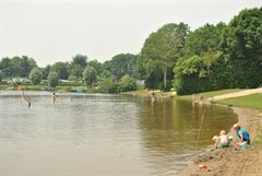 Zwemwater met meerdere kinderen en volwassenen in het water. Op het strandje aan de rechterkant zijn twee kinderen met zand aan het spelen.