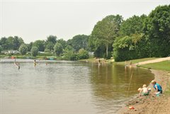 Zwemwater met meerdere kinderen en volwassenen in het water. Op het strandje aan de rechterkant zijn twee kinderen met zand aan het spelen.