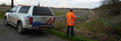 Medewerker van het waterschap controleert de watergang. Links staat een auto van het waterschap.