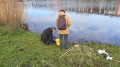 Twee personen meten de waterkwaliteit bij een sloot met een emmer, meethark, secchi-schijf en een schepnet